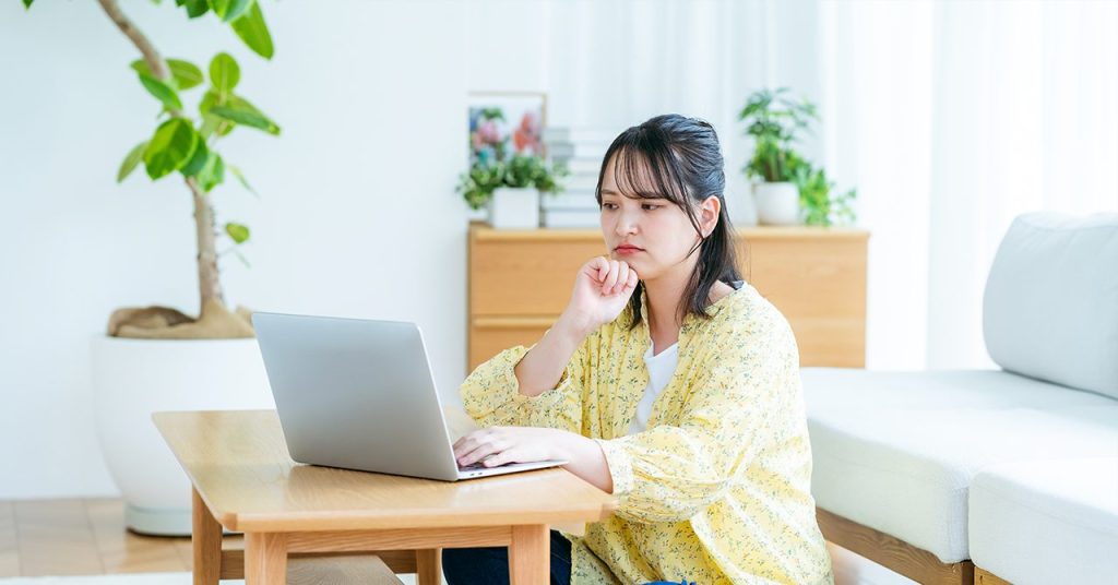 明るい室内で座卓に向かい、手元のノートパソコンを見ながら考え込む若い女性。