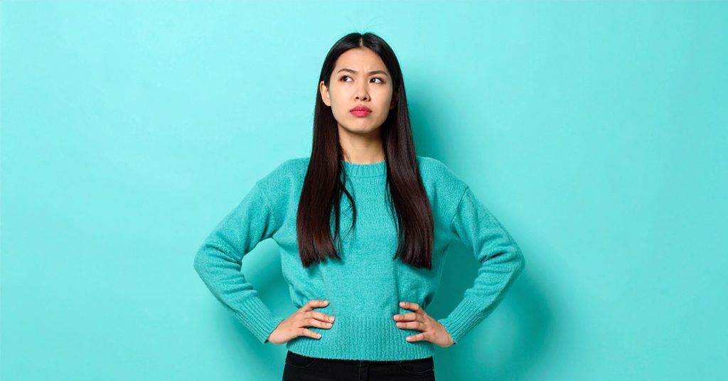 疑いの表情を見せる女性のビジュアル。両手を腰にあて、目線は空を睨んでいる険しい表情。